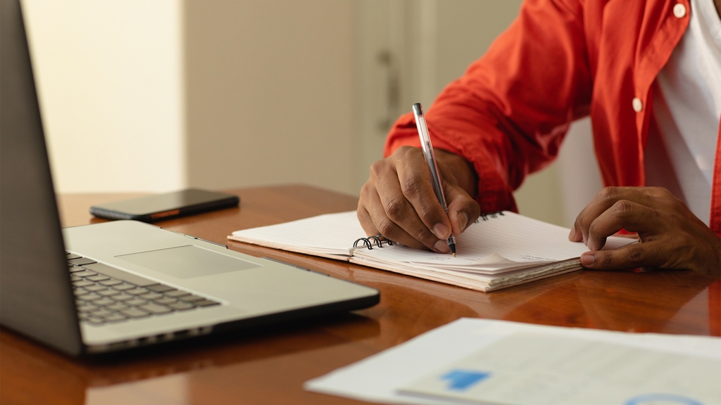 Pessoa escrevendo em um caderno sobre uma mesa, com um notebook aberto ao fundo. 