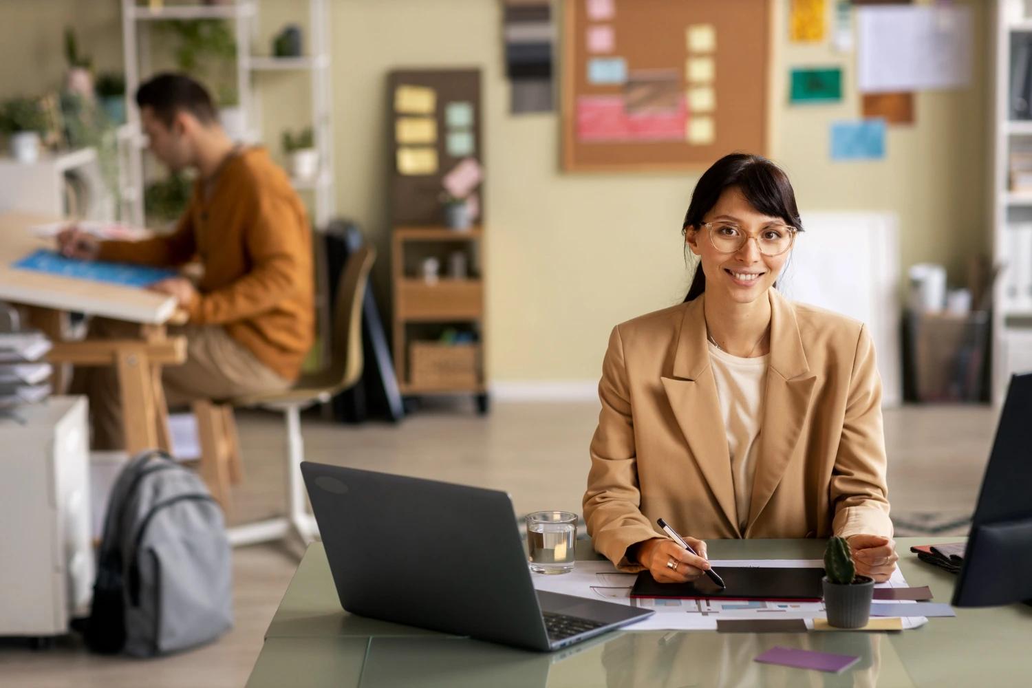 Mulher sorridente trabalha em sua mesa com laptop e tablet, enquanto um colega trabalha ao fundo, ilustrando a rotina da carreira executiva.