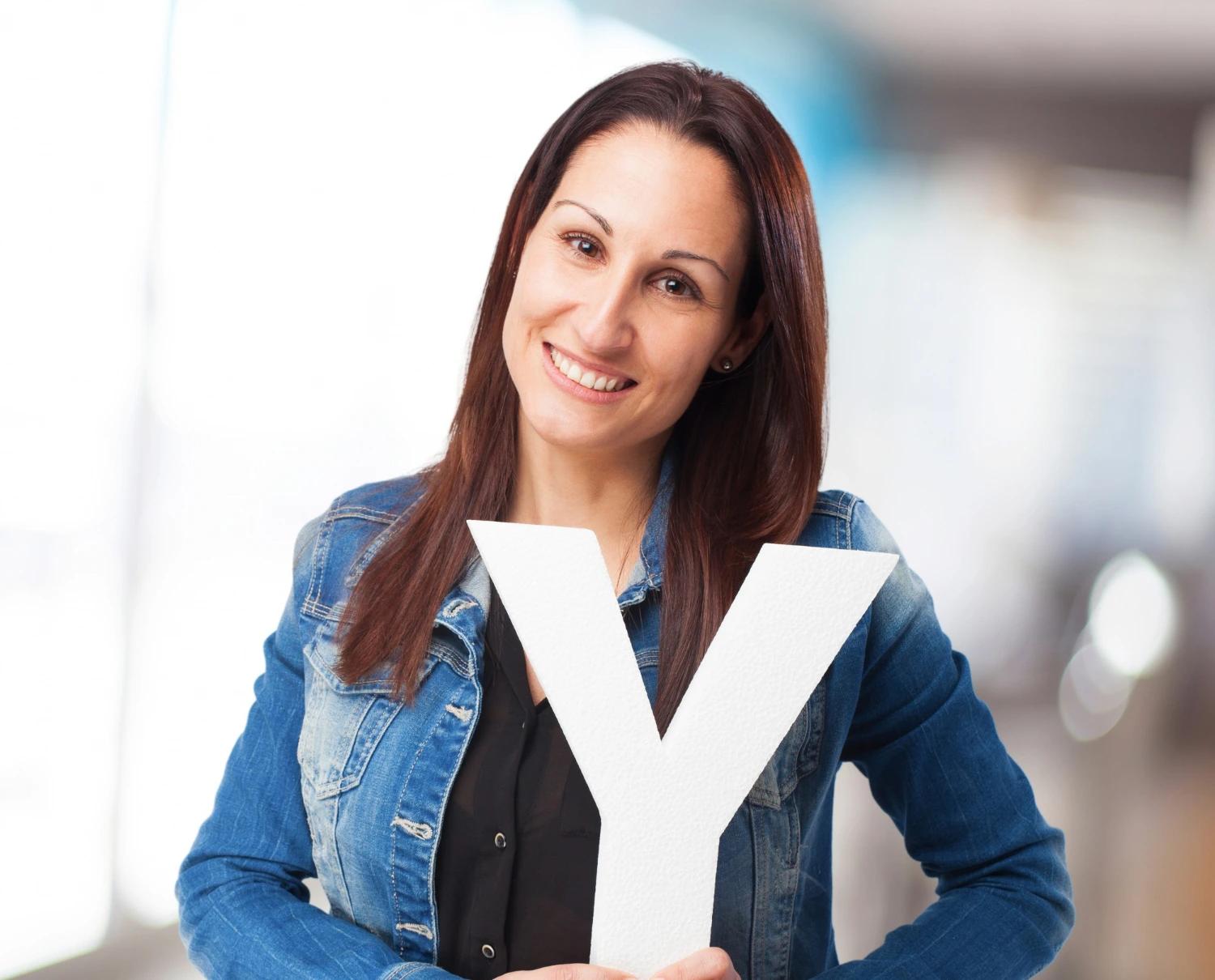 Uma mulher de cabelos lisos castanhos, sorridente, está em pé em um ambiente interno desfocado ao fundo. Ela veste uma jaqueta jeans azul sobre uma blusa preta. Segura com as duas mãos uma grande letra branca em formato de "Y", posicionada à frente do corpo, ilustrando o formato da carreira em Y.