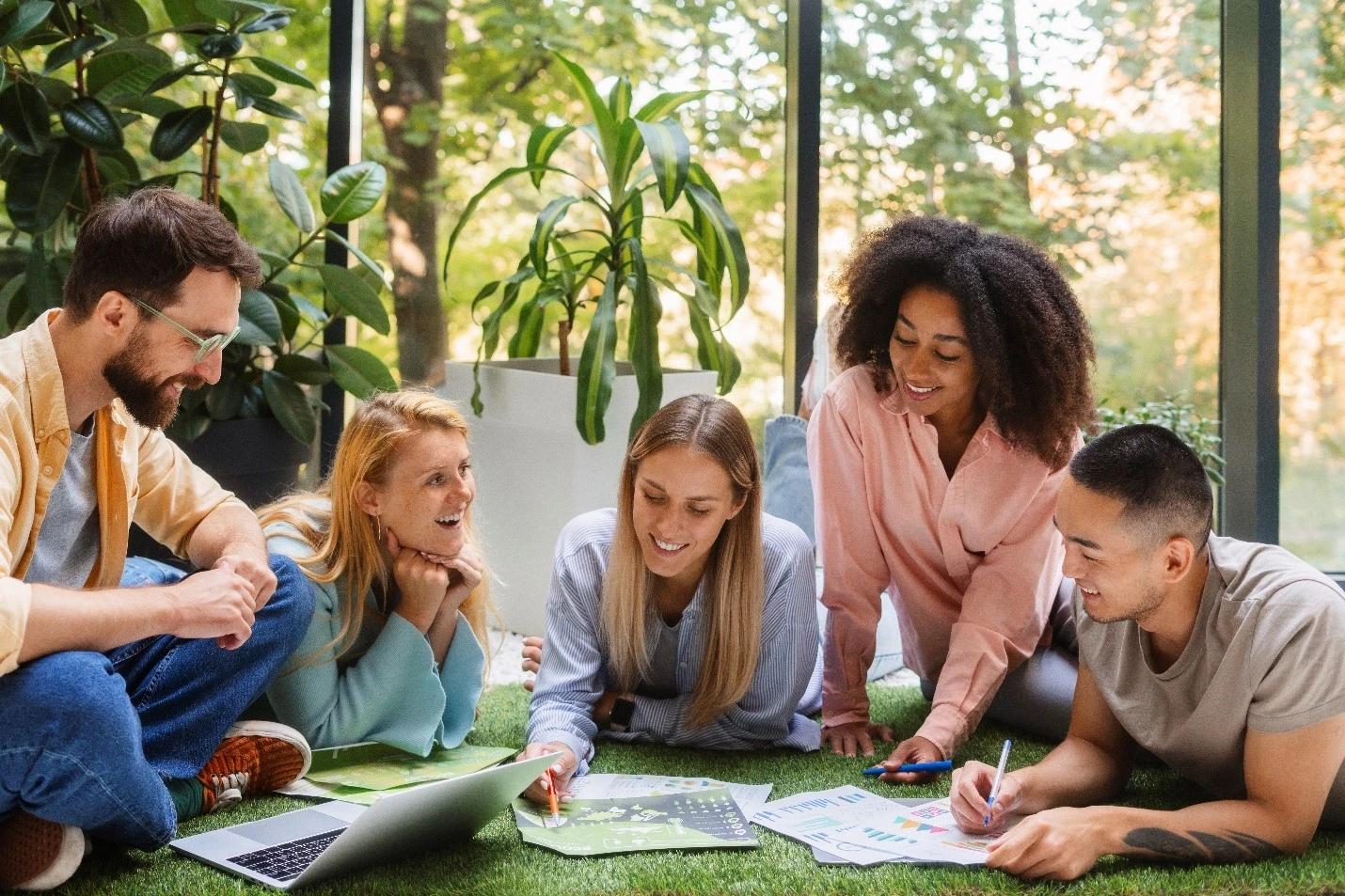 Cinco pessoas jovens, de diferentes perfis, estão sentadas no chão sobre grama sintética, cercadas por plantas e janelas envidraçadas. Elas analisam relatórios coloridos e um notebook, discutindo soluções sustentáveis em clima descontraído. A foto ilustra diversidade, colaboração e consciência ambiental, valores que são aprofundados em cursos de ESG.