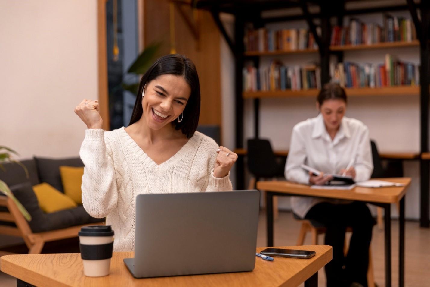 Uma mulher comemora sorridente diante de um notebook em um ambiente de estudo, demonstrando satisfação com seus resultados. A cena representa a realização profissional alcançada por meio de cursos para educadores, que fortalecem a confiança e o aprimoramento contínuo na carreira docente.