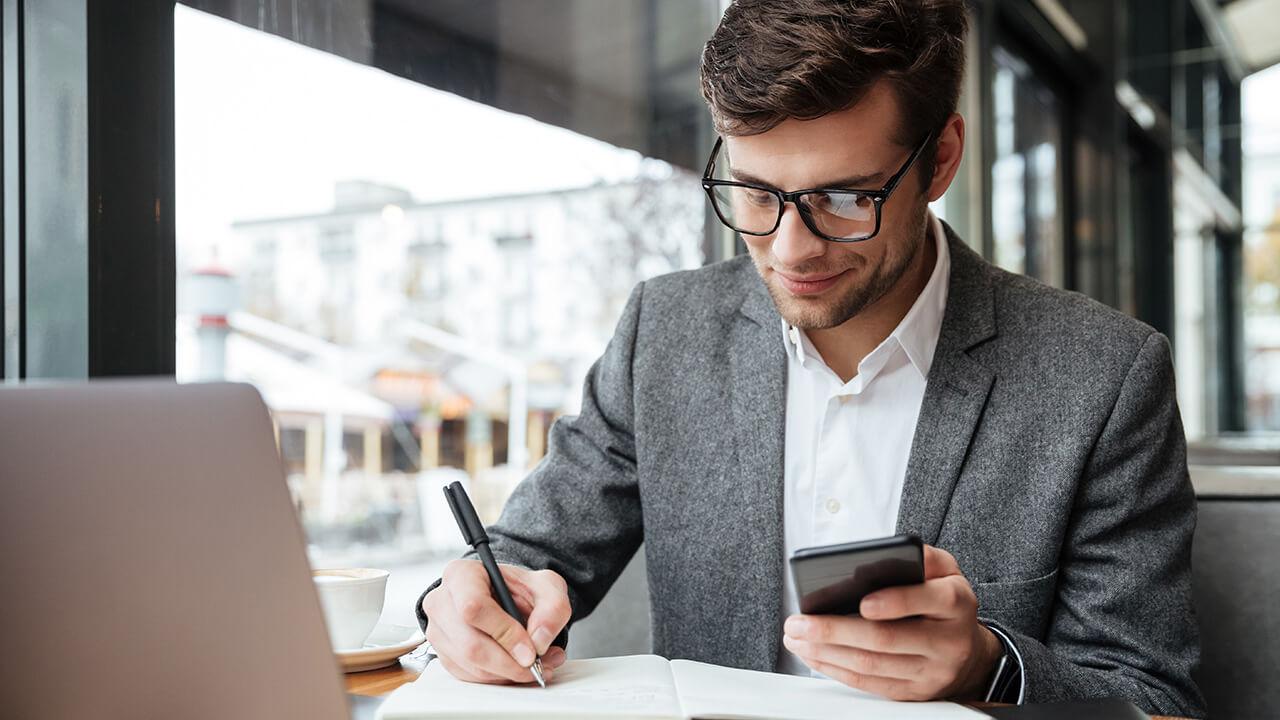 Homem em frente a um computador com calculadora e caderno não mão, enquanto faz alguns cálculos no papel.