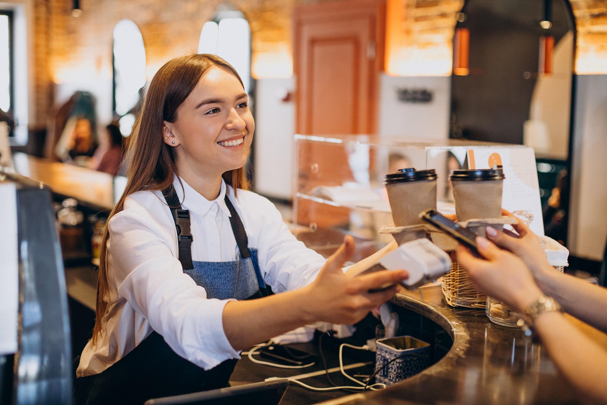 Atendente sorridente realizando um pagamento no caixa de uma cafeteria, demonstrando as habilidades de um operador de loja, como atendimento ao cliente, agilidade e simpatia.