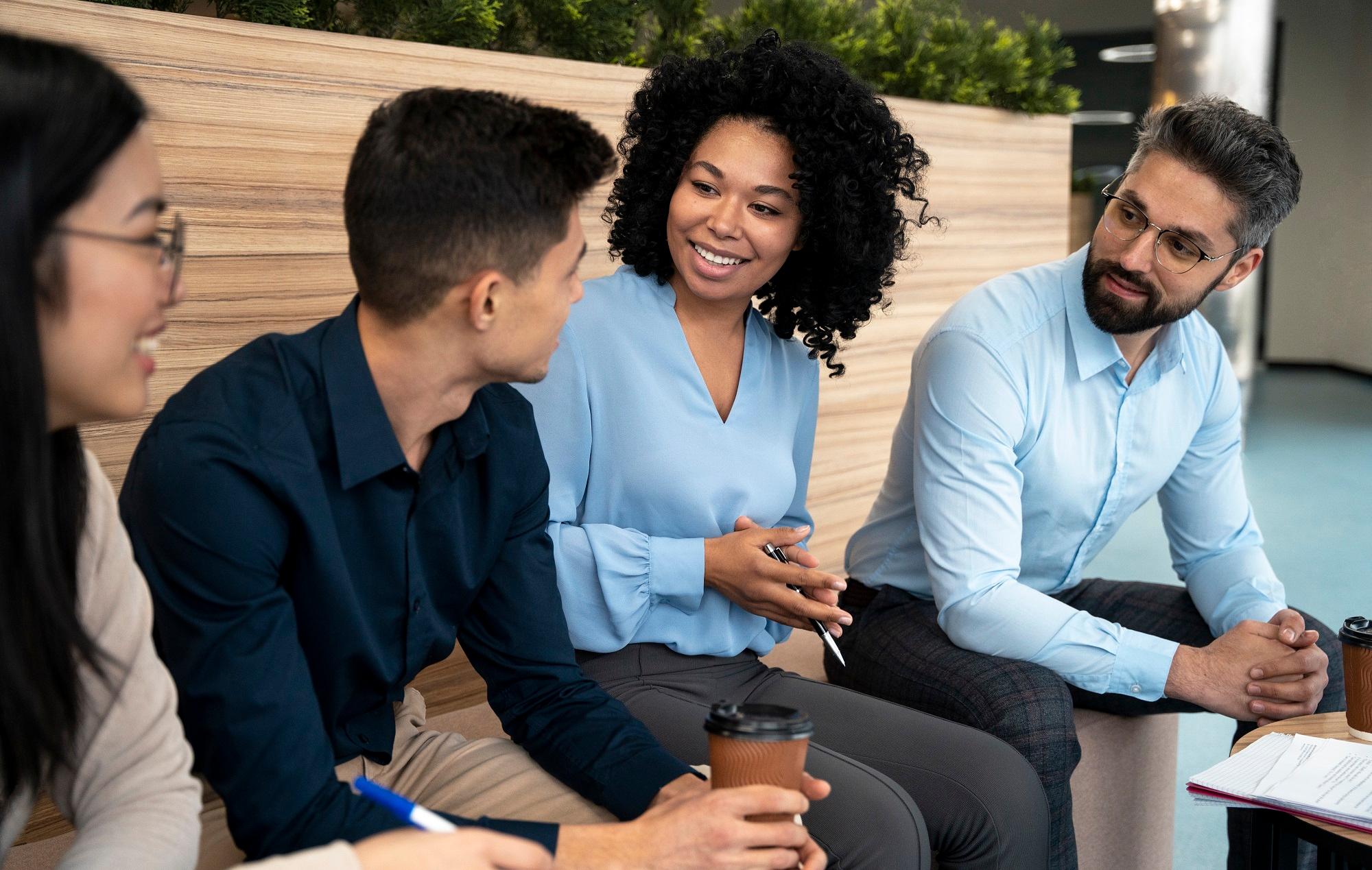 Grupo de quatro profissionais conversando de forma descontraída, sorrindo e trocando ideias, representando a importância da comunicação interpessoal no ambiente de trabalho.