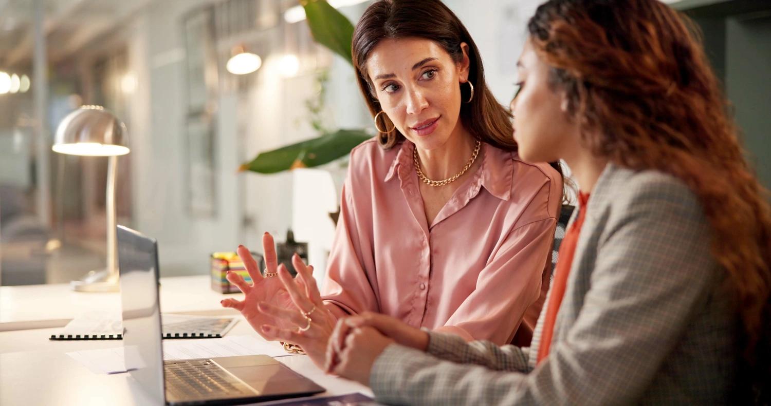 Uma gerente passando um feedback negativo para uma colaboradora, em sala de trabalho, com laptop na mesa.